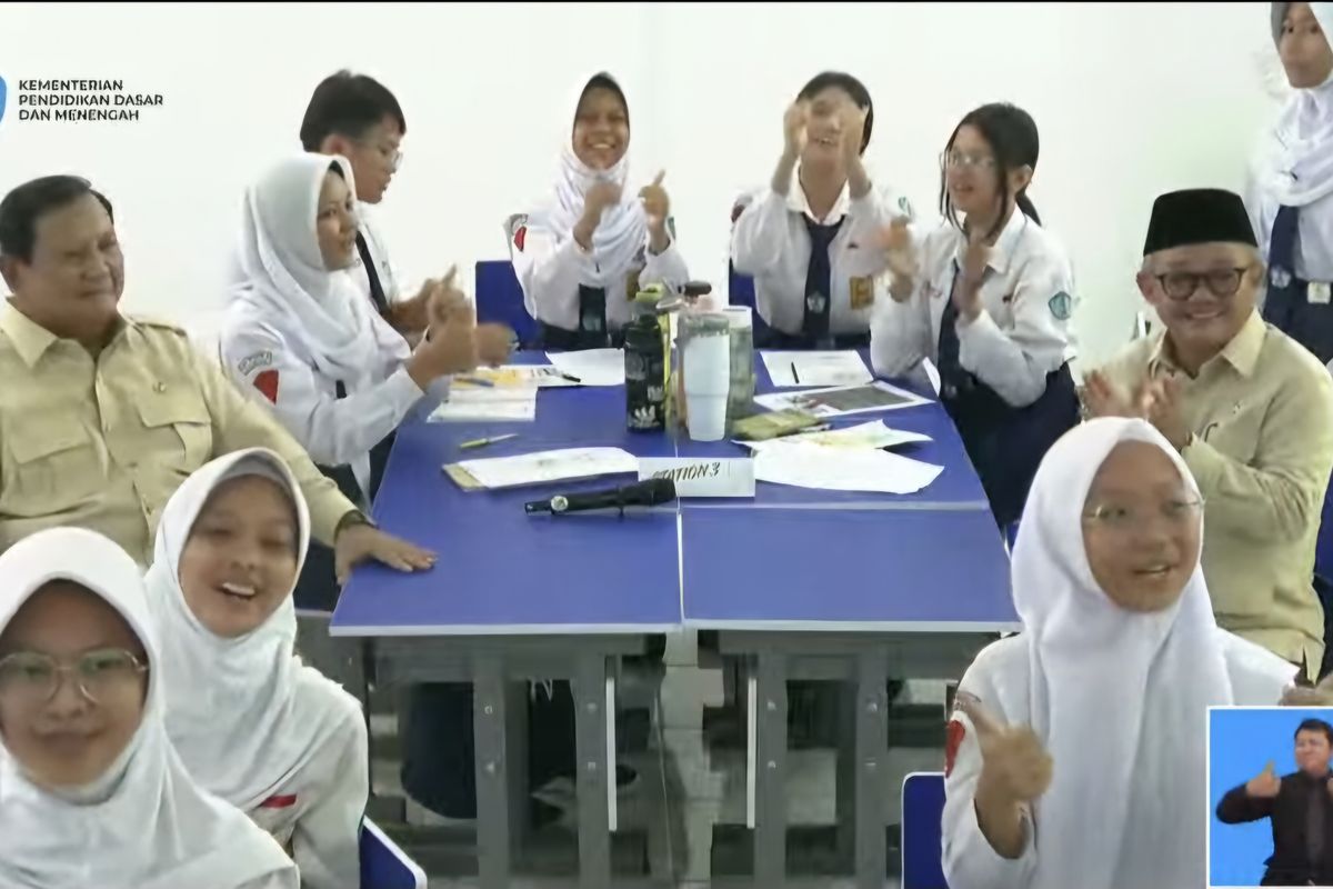 Sejumlah siswa SMP mengenakan seragam putih duduk bersama di sebuah meja kelas sambil tertawa dan bertepuk tangan, didampingi oleh pejabat Kementerian Pendidikan Dasar dan Menengah saat kunjungan program Interactive Flat Panel.