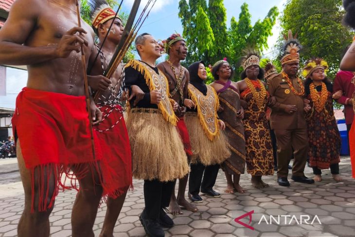Sekelompok pria dan wanita mengenakan pakaian adat Papua, beberapa membawa busur dan memakai hiasan bulu, berdiri berjejer sambil menari di luar ruangan.