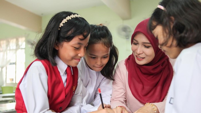 Seorang guru berhijab merah membimbing tiga siswa sekolah dasar yang sedang menulis bersama di kelas dengan suasana hangat dan fokus.