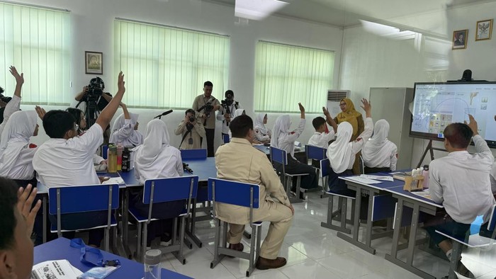 Suasana kelas di SMPN 4 Kota Bekasi: para siswa berseragam putih banyak yang mengangkat tangan, guru berhijab kuning berdiri di dekat smartboard/layar interaktif, sementara beberapa jurnalis memotret dari belakang kelas.