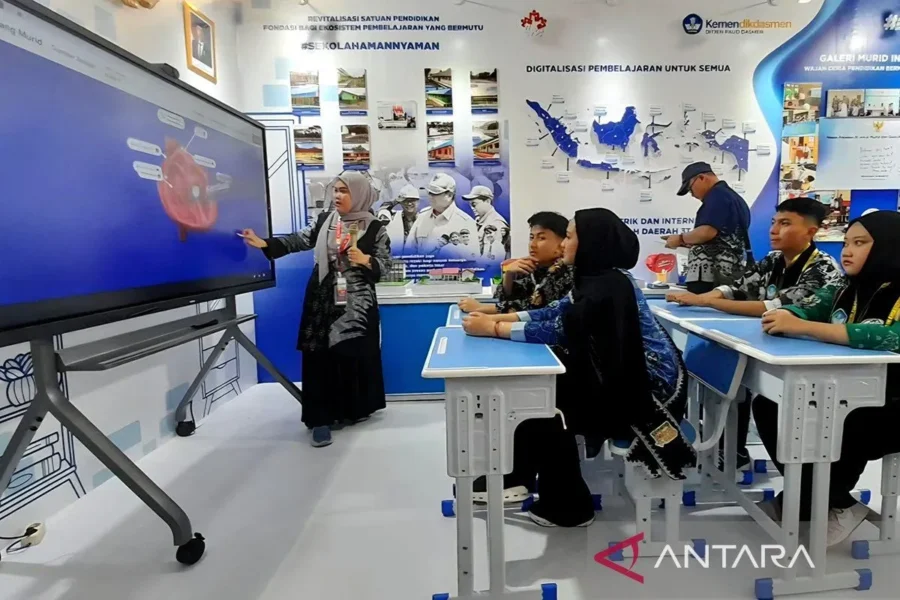 Seorang guru mengajar menggunakan papan interaktif digital di depan siswa yang duduk di kelas dan memperhatikan pelajaran.